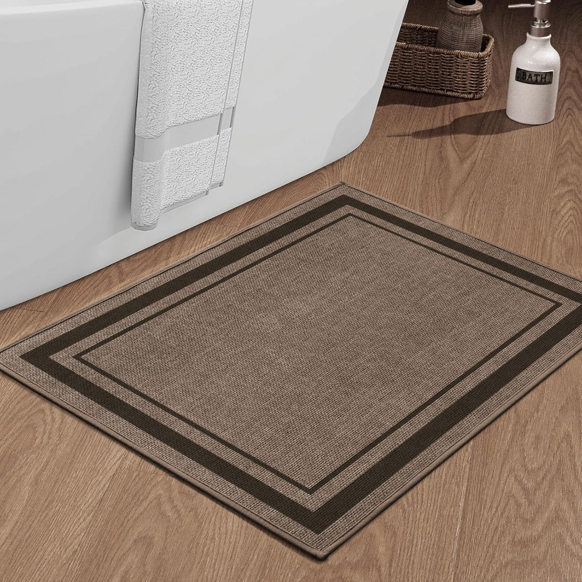 Close-up of a square beige bath rug with textured weave and dark border on wood floor