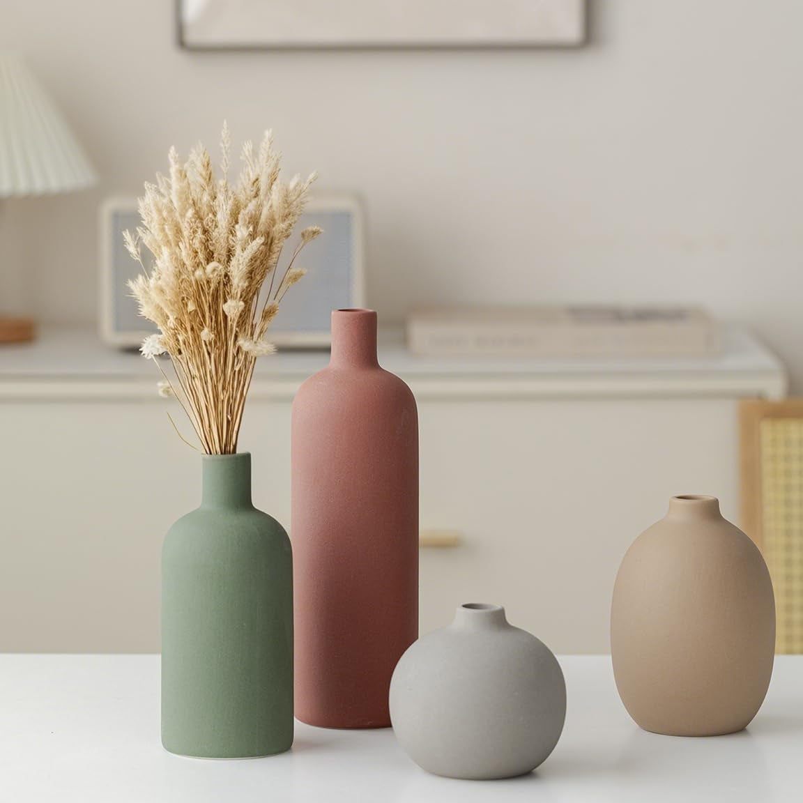 Four matte vases on a tabletop with tall terracotta bottle in front of a blurred sideboard and framed art
