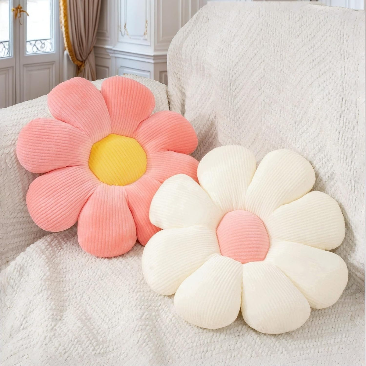 Angled close of a coral-pink daisy cushion with a yellow center beside a cream daisy, resting on a knitted throw.