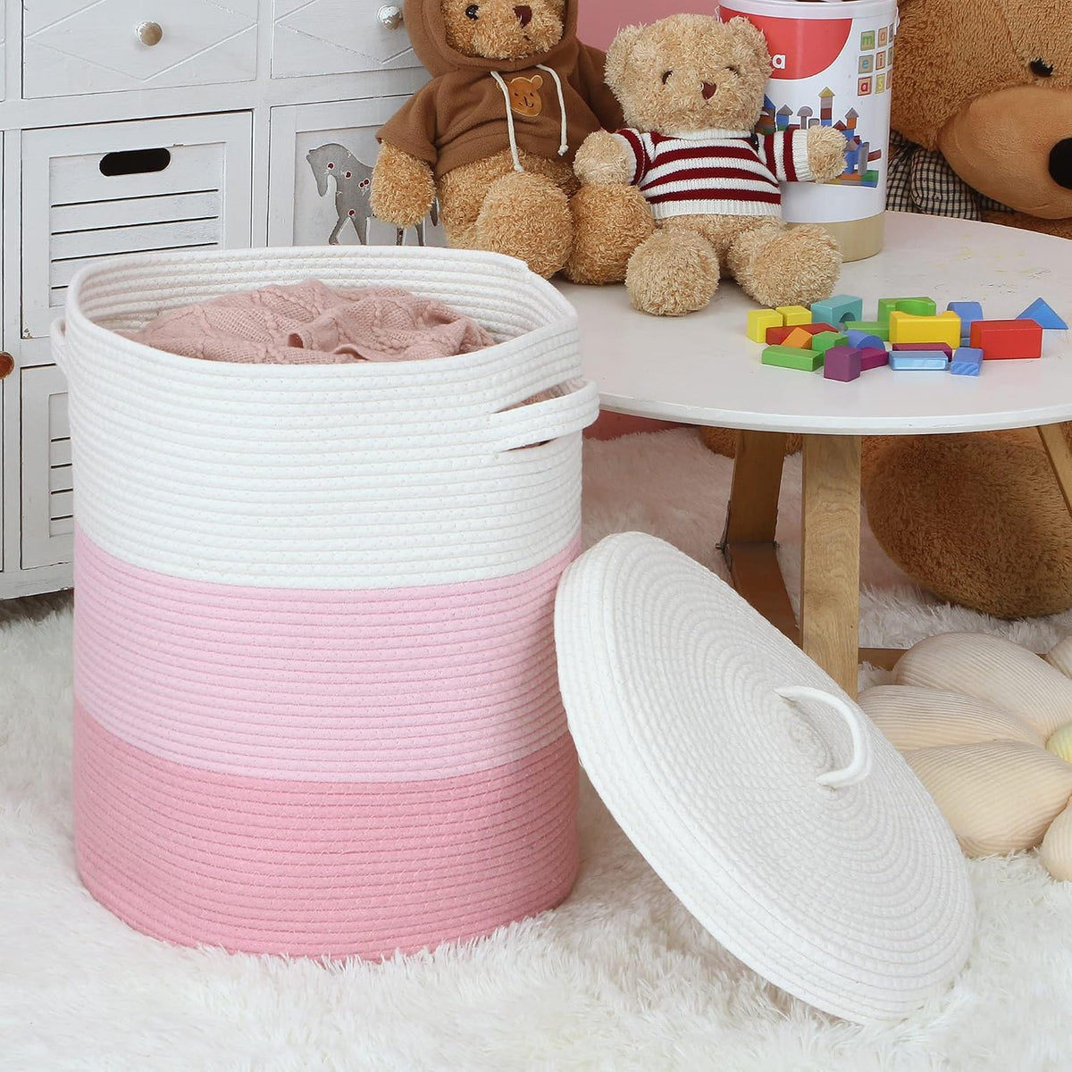 Pink and white woven rope laundry basket with the lid removed beside a low play table and stuffed toys