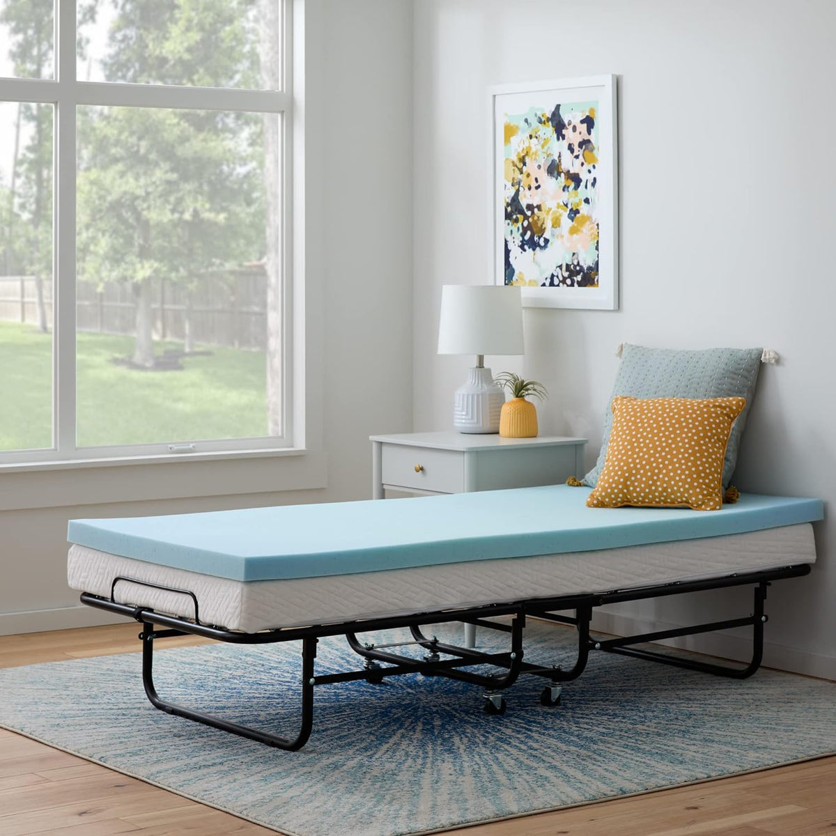 Fold-out metal guest bed with a light blue foam topper, white mattress, bedside table, lamp and decorative pillows.