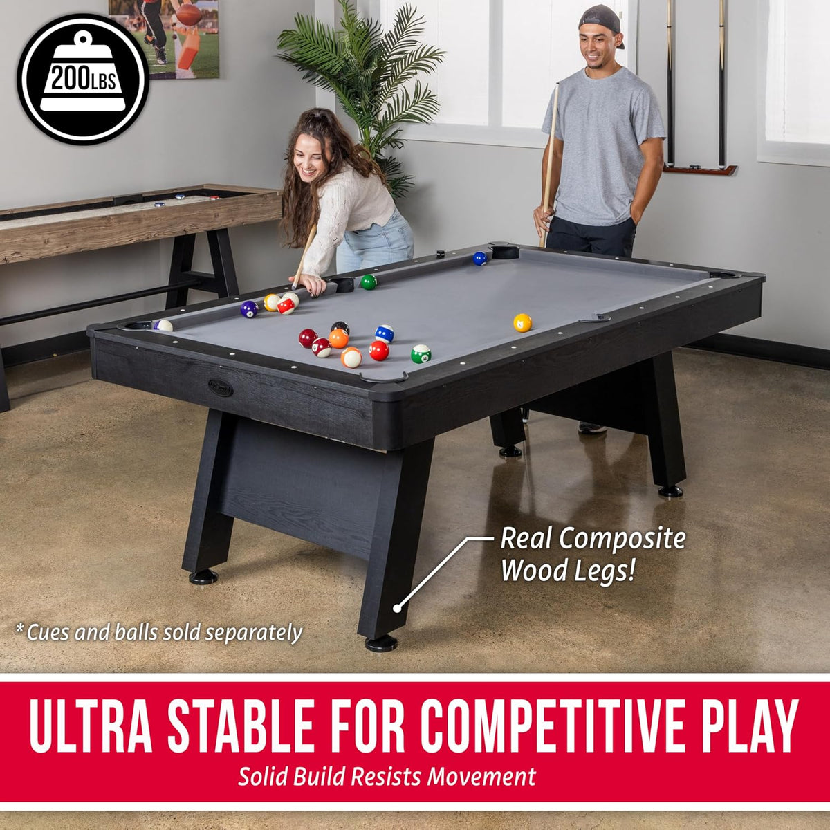 Two people playing on a gray-felt pool table in a game room, balls scattered across the table