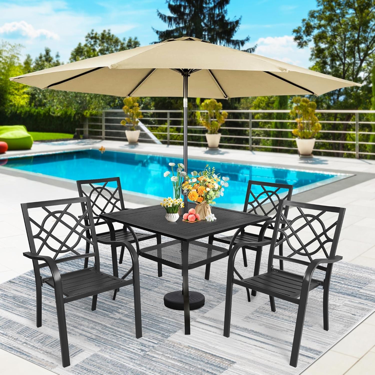 Square black table and four chairs on a striped outdoor rug by a pool, topped with a bouquet and a beige umbrella overhead.