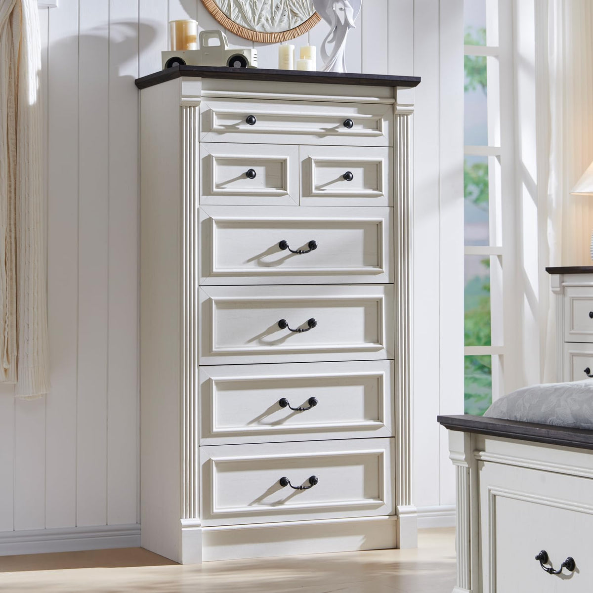Angled lifestyle view of the white tall dresser with dark tabletop shown beside a bed in a styled room