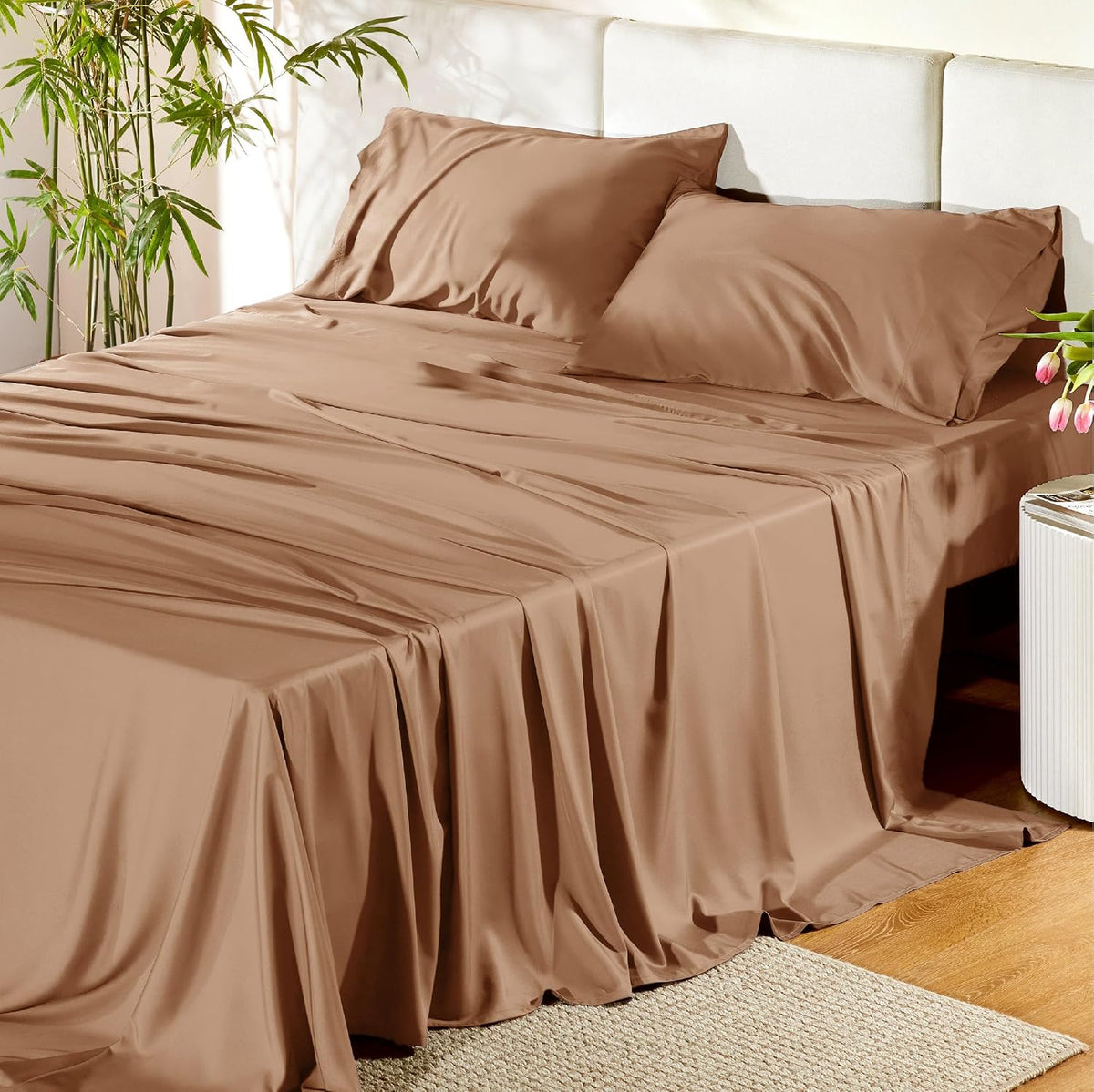 Front-facing bed with warm chestnut-brown sheets draped to the floor and a potted bamboo plant nearby.