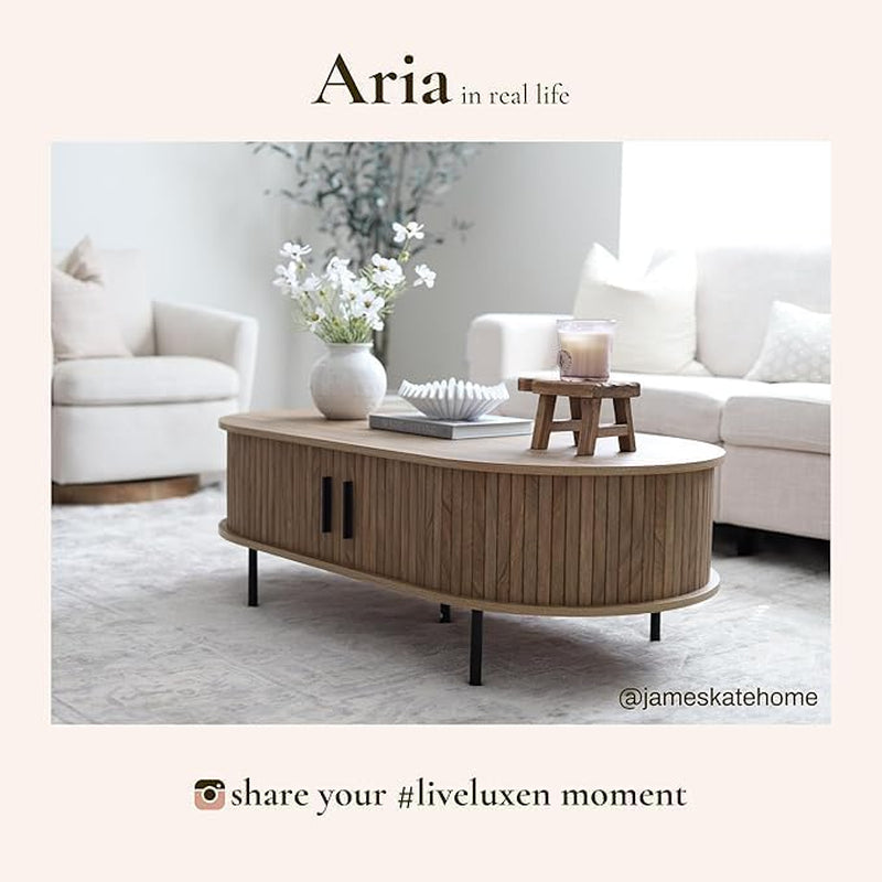 Oval oak-grain coffee table with closed tambour doors, small wooden stool and vase on top, light sofas behind.