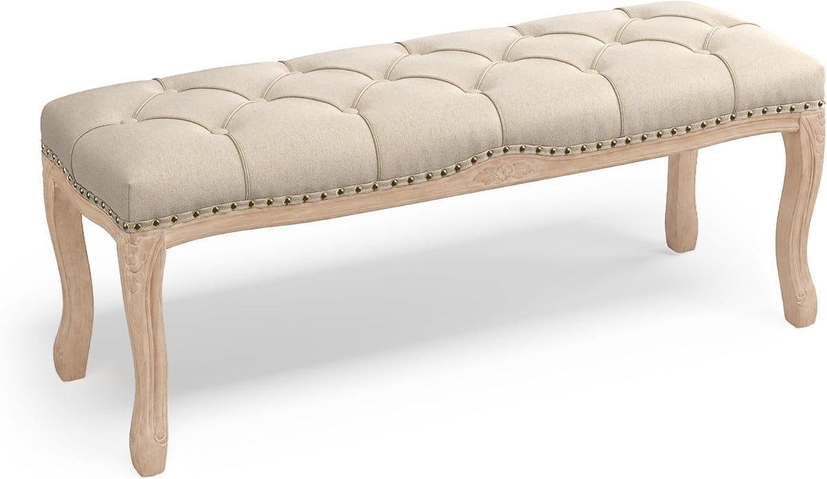 Studio shot of a beige tufted bench from a low angled perspective showing carved pale wood legs and nailhead trim.