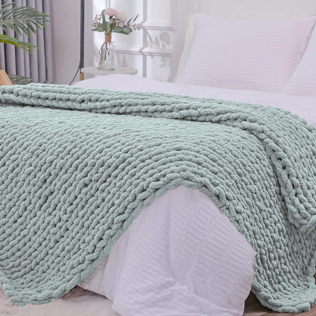 Sage-green chunky knit throw folded over the edge of a pale pink bedspread showing braided rows.