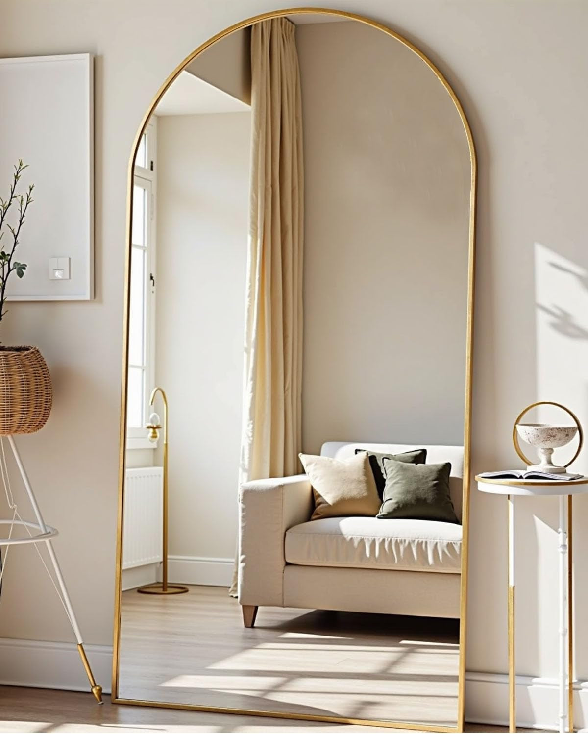 Arched floor mirror with slim gold frame leaning against a wall reflecting a sunlit sofa and window.