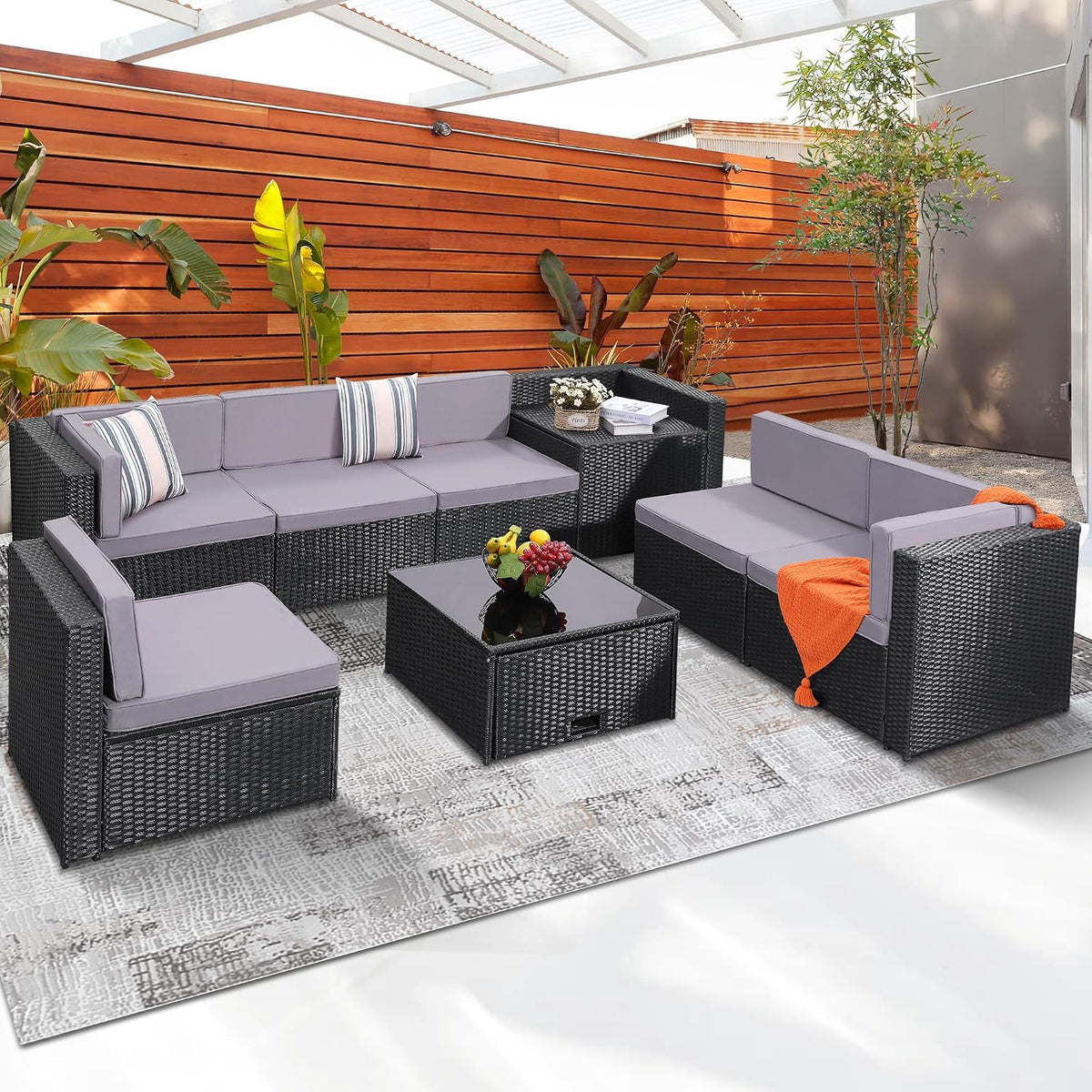 Modular wicker sofa set arranged under a pergola with gray cushions, coffee table, plants and wood fence.