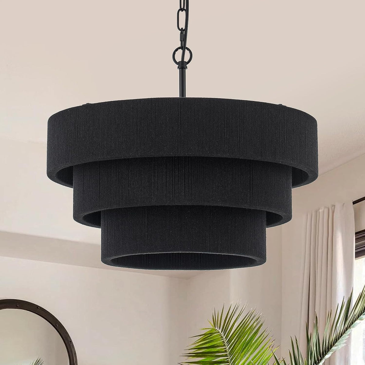 Black three-tiered drum pendant light suspended by a black chain in a bright room with curtains, potted palm, and round