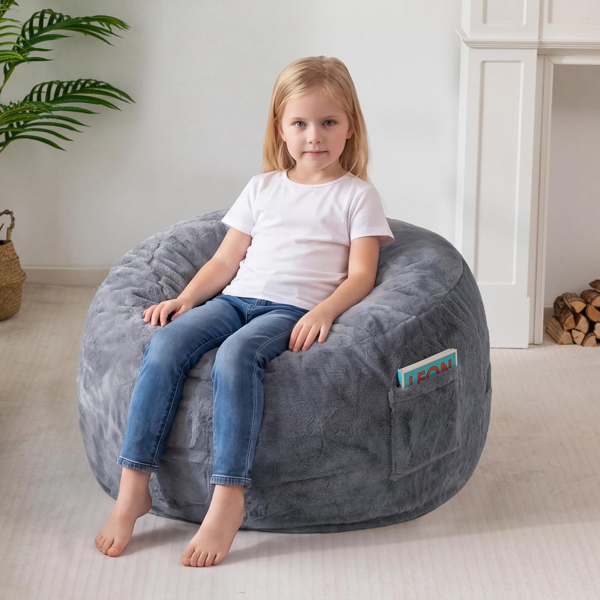 Young girl sitting upright with legs dangling on a gray bean bag; a colorful book peeks from the side pocket.