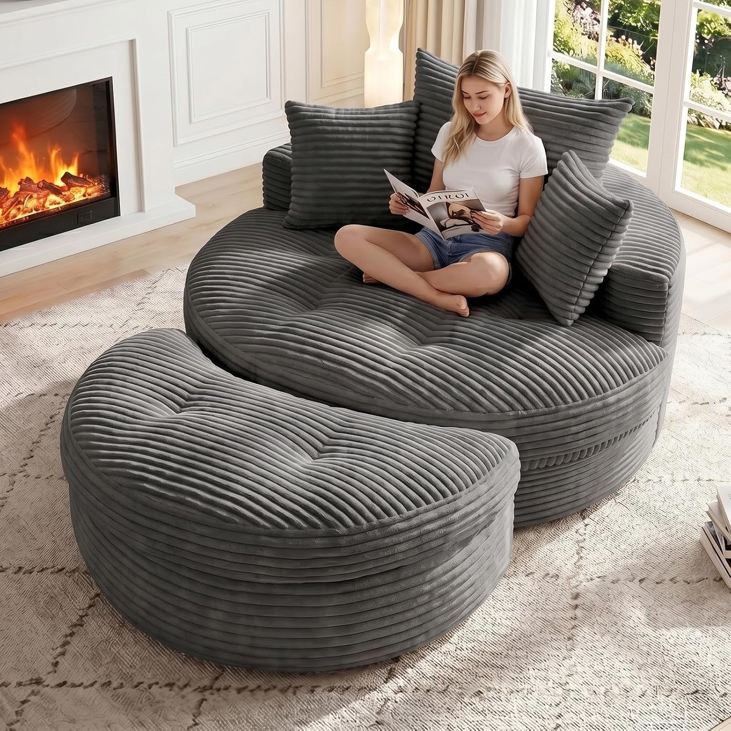 Round oversized corduroy floor chaise with matching crescent ottoman in charcoal gray, woman reading by a fireplace