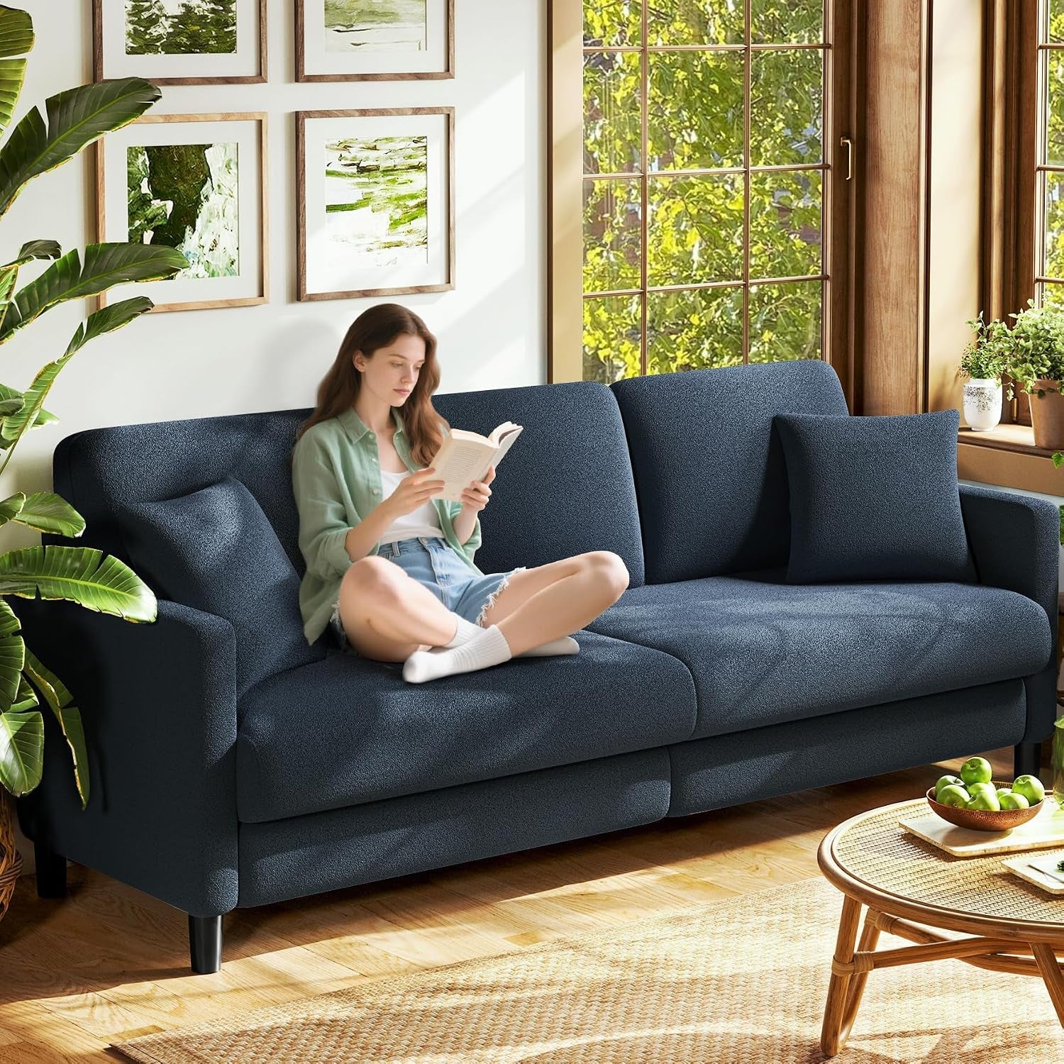 Woman reading on a dark blue sofa set by a sunlit window with matching pillow