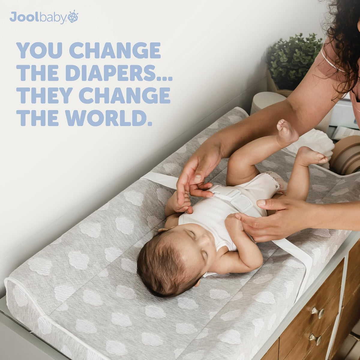 Caregiver fastening a white safety strap as a baby lies on the gray patterned contoured changing pad on a dresser.