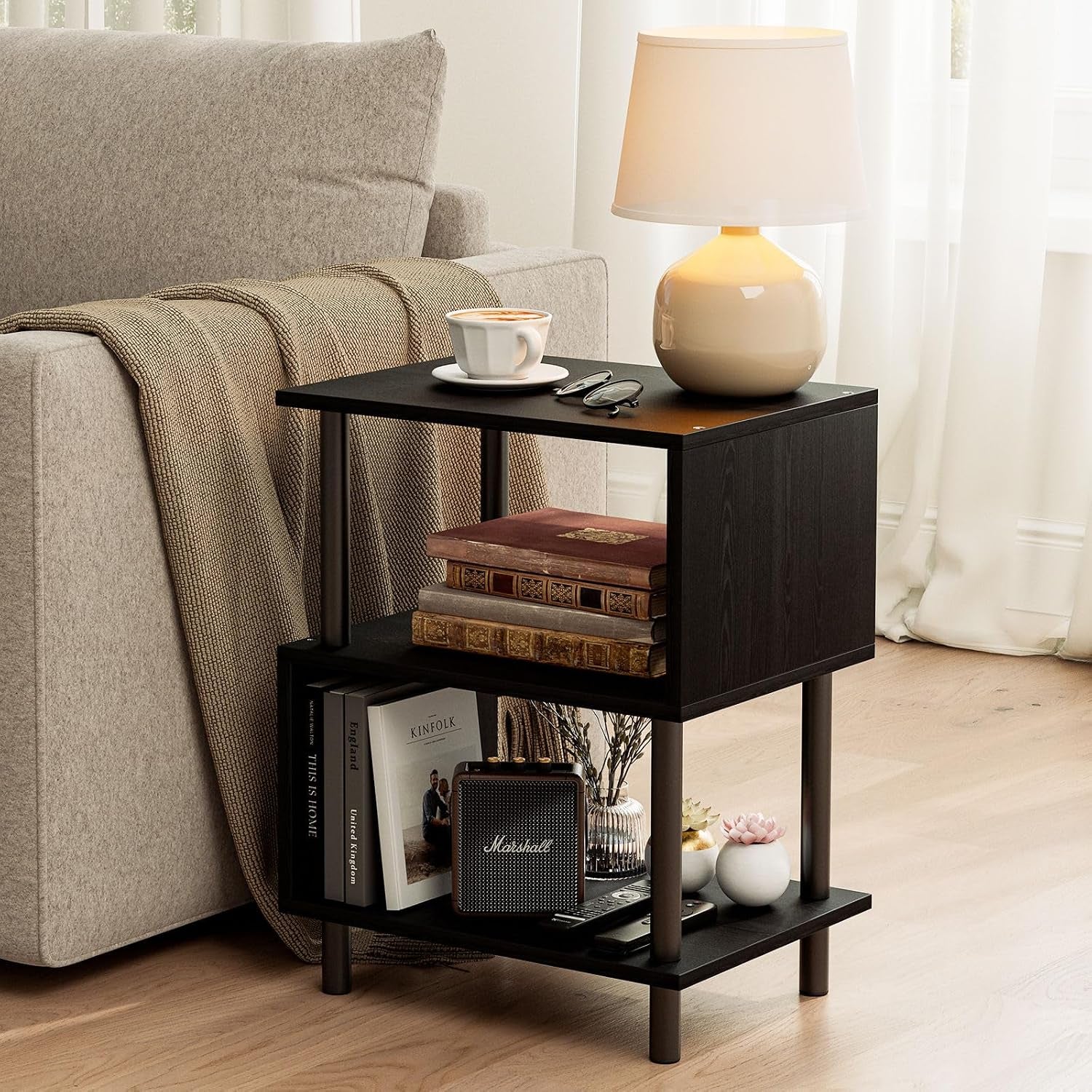 Black three-tier side table next to sofa with lamp, stacked books and decorative vases