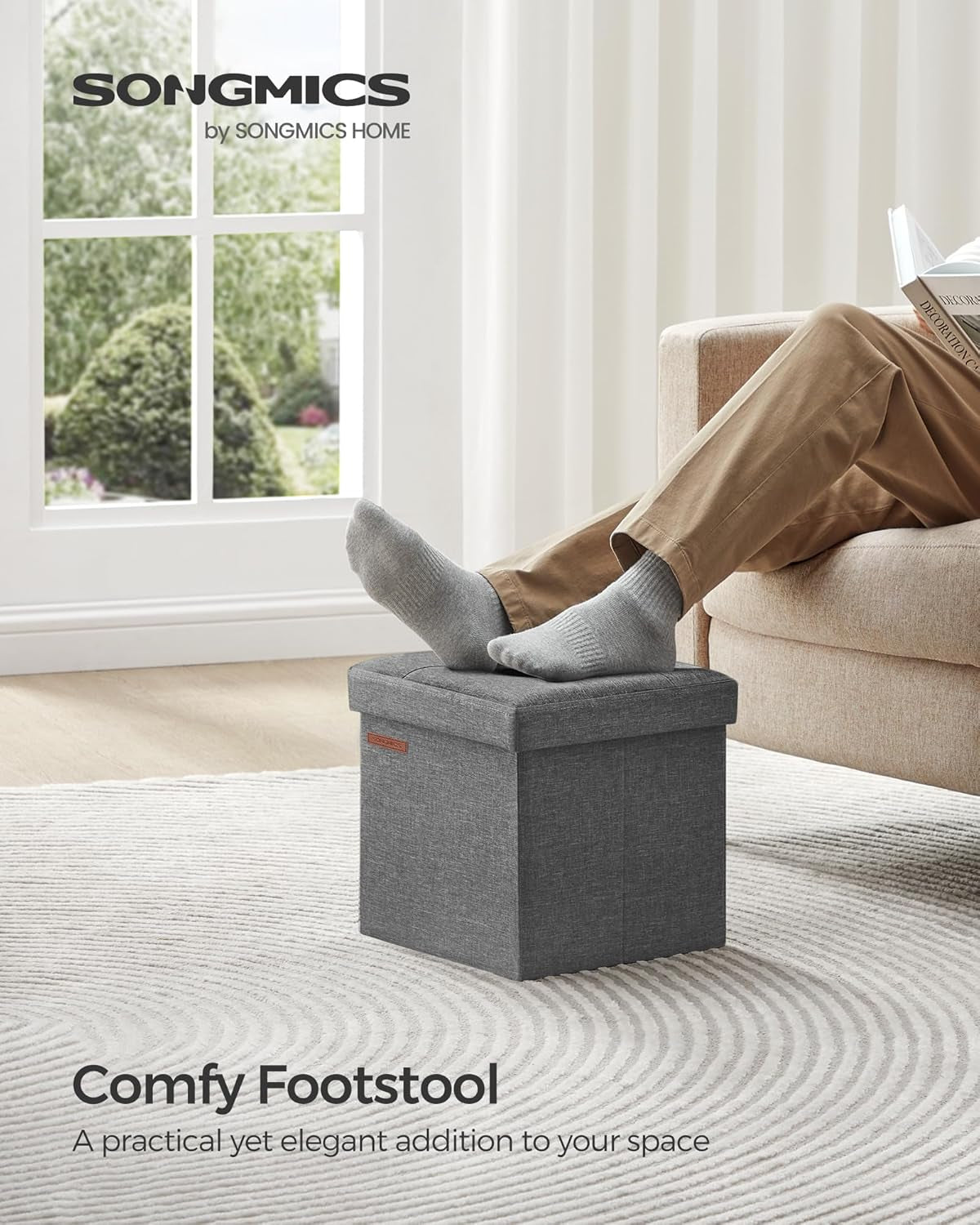 Living room scene with a person resting socked feet on a gray storage ottoman next to a beige sofa by a window.