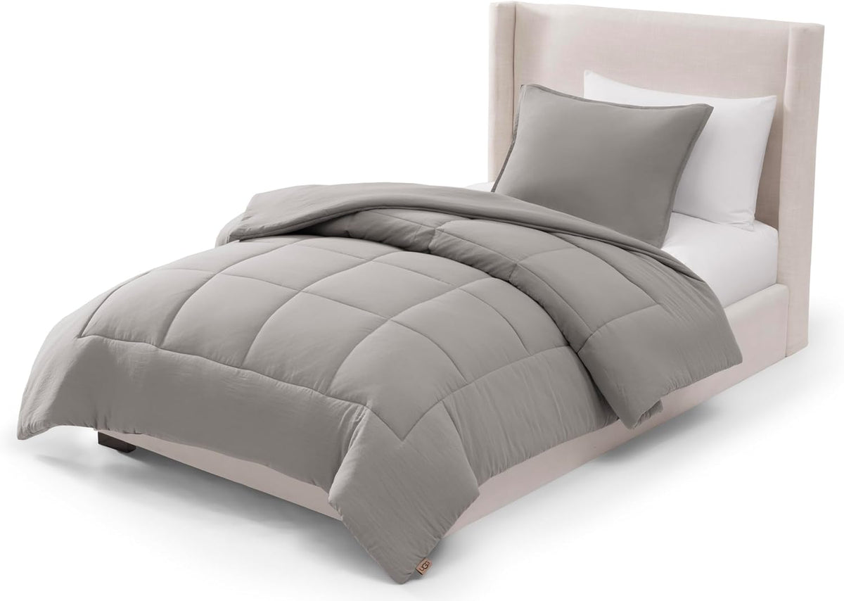 Full/queen bed with a lightly folded pale grey box‑stitched comforter and one matching sham propped against a beige