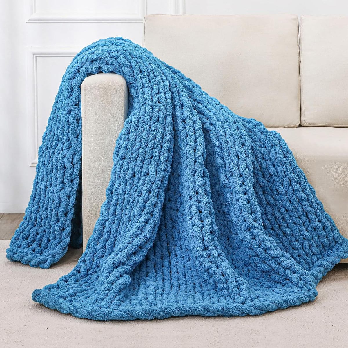 Vivid blue chunky knit blanket draped over a sofa arm, showing long braided folds that reach the floor.