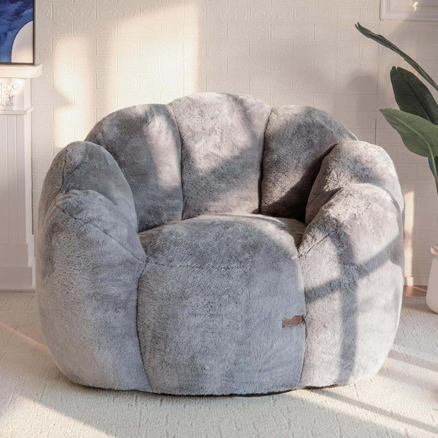 Plush floral-shaped bean bag chair in light gray placed in a bright room with a potted plant at right.