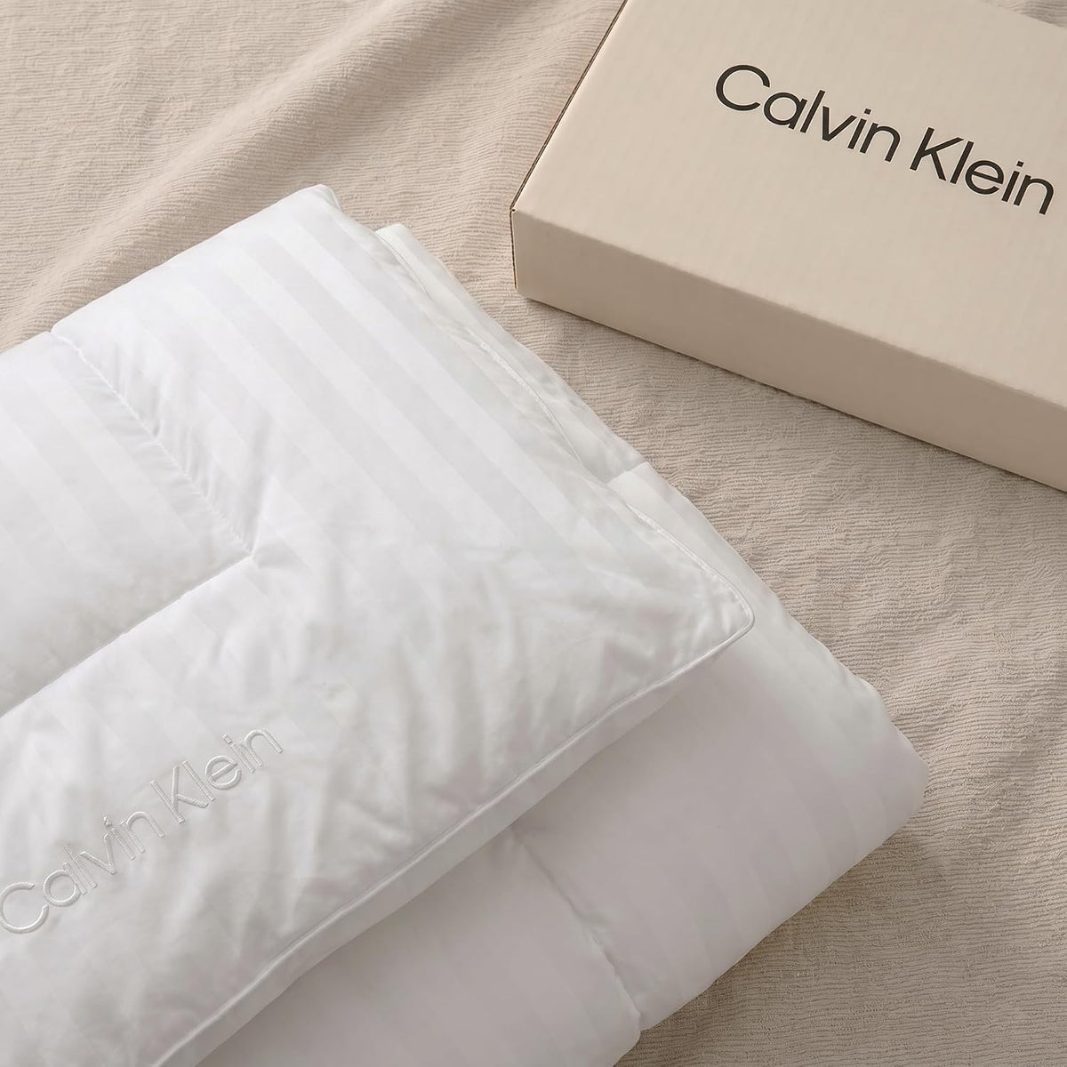 Top-down view of a folded white striped comforter and matching pillow on a textured beige surface with a beige box nearby
