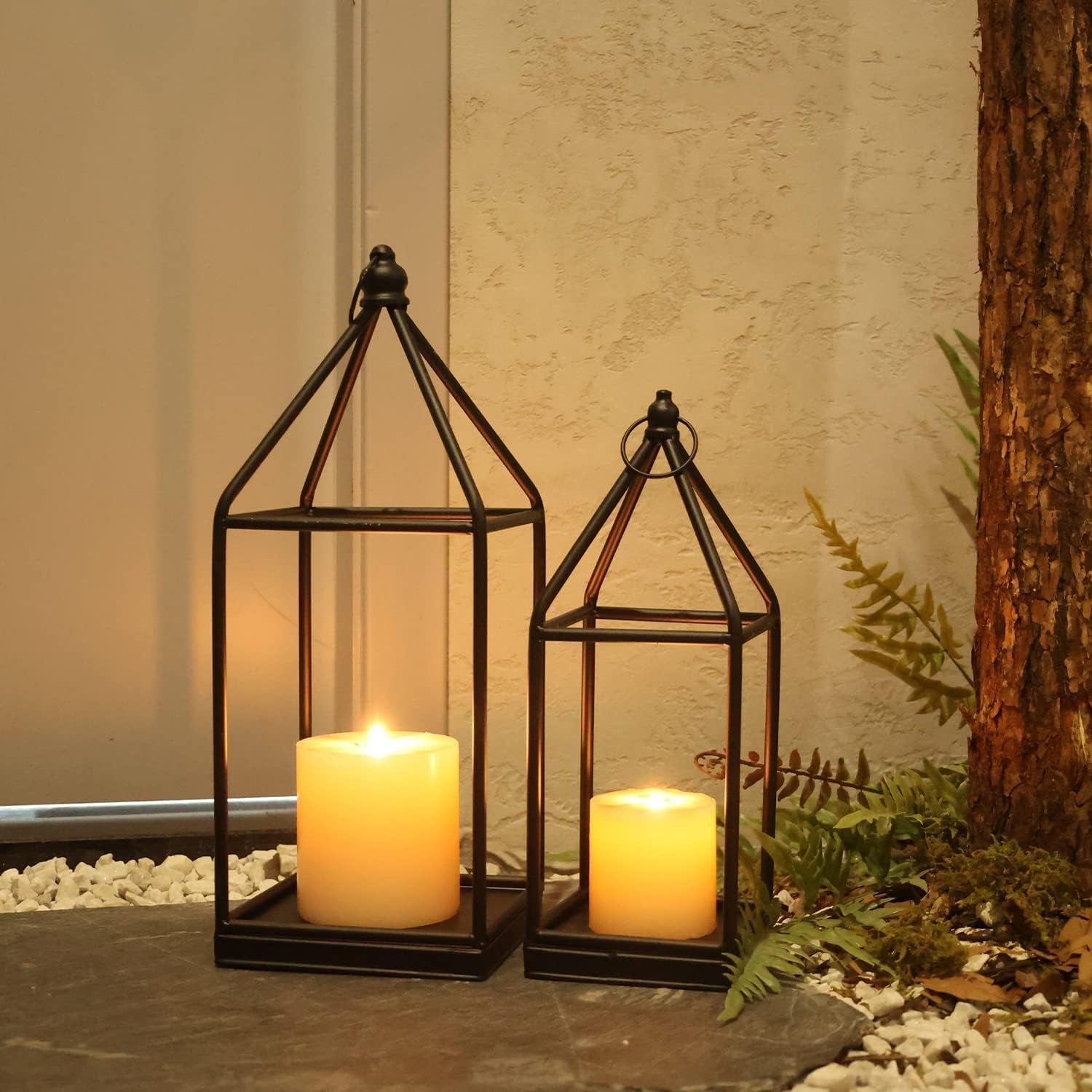 Close-up of two lanterns with warm flickering candles beside a tree and low groundcover stones.