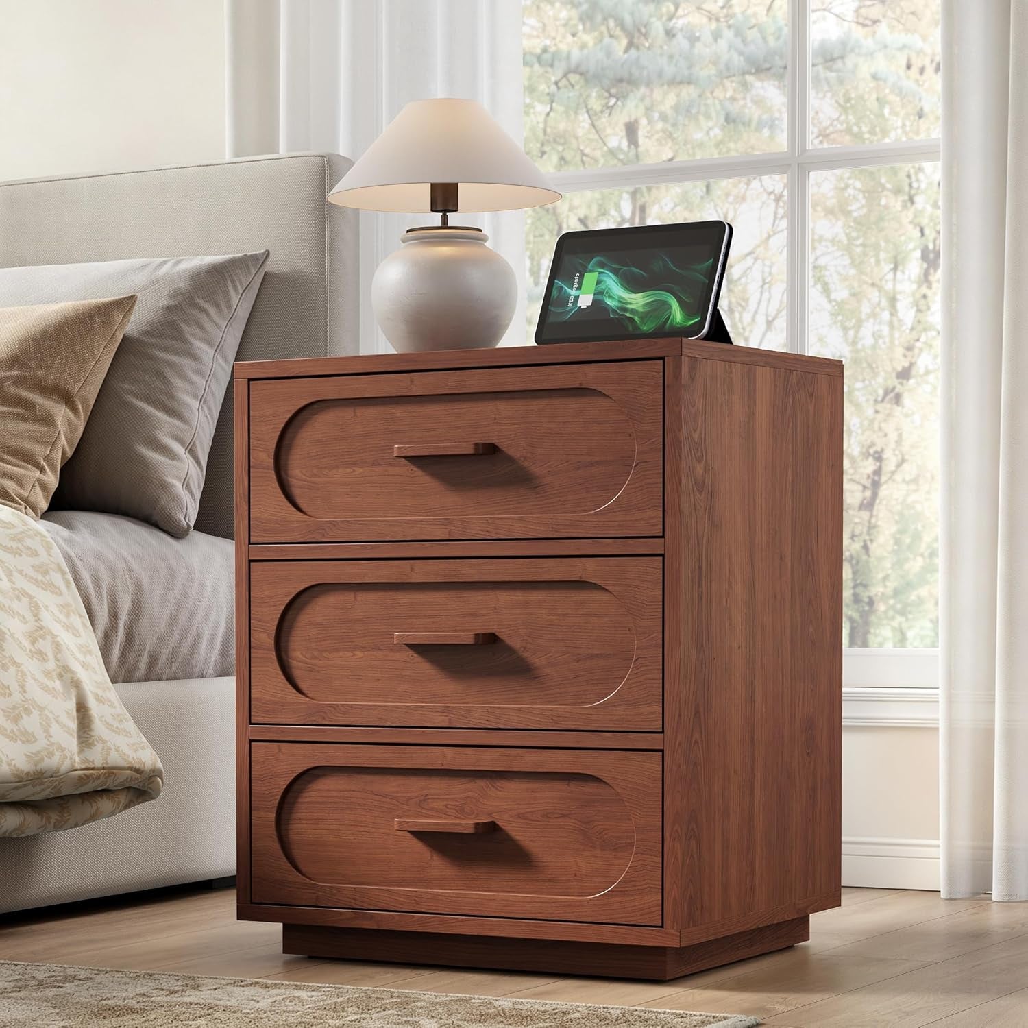 Dark-brown three-drawer nightstand next to a bed topped with a lamp and tablet