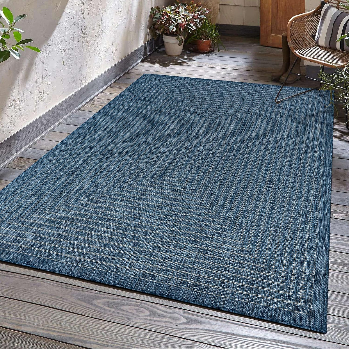 Angled view of a blue-green woven outdoor rug on a wooden porch with potted plants and a chair.
