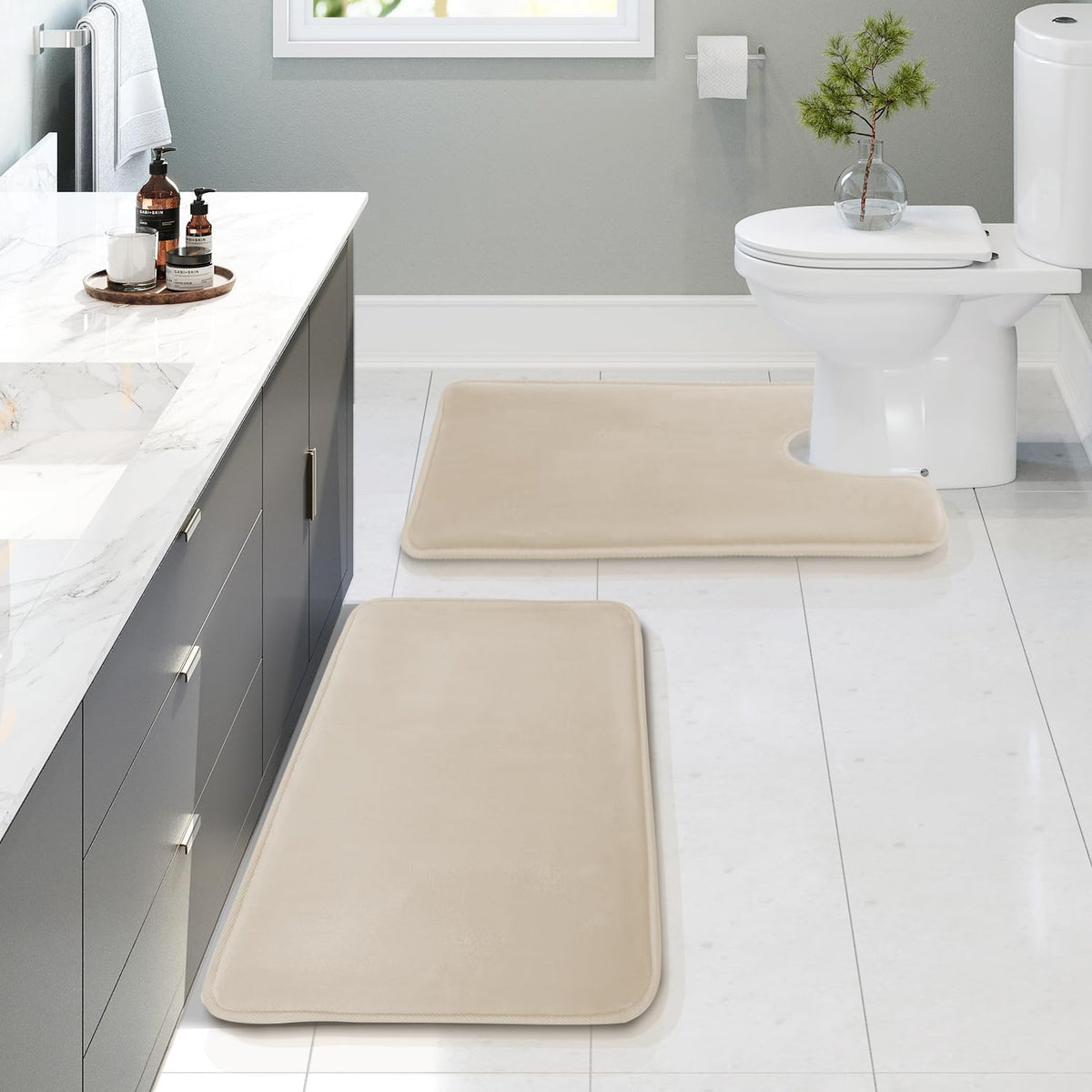 Beige three‑piece bath rug set arranged on a white tiled bathroom floor near a toilet and vanity.