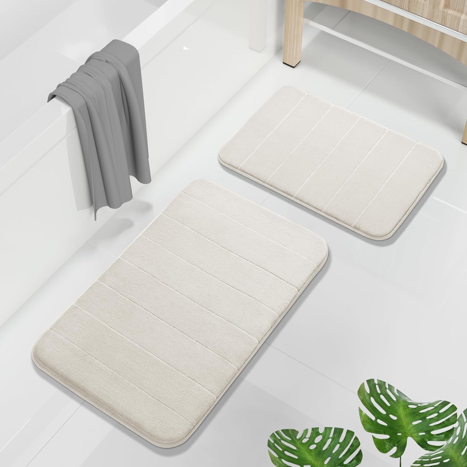 Beige memory-foam bath mats beside a bathtub with a gray towel draped over the tub and green plant leaves nearby.