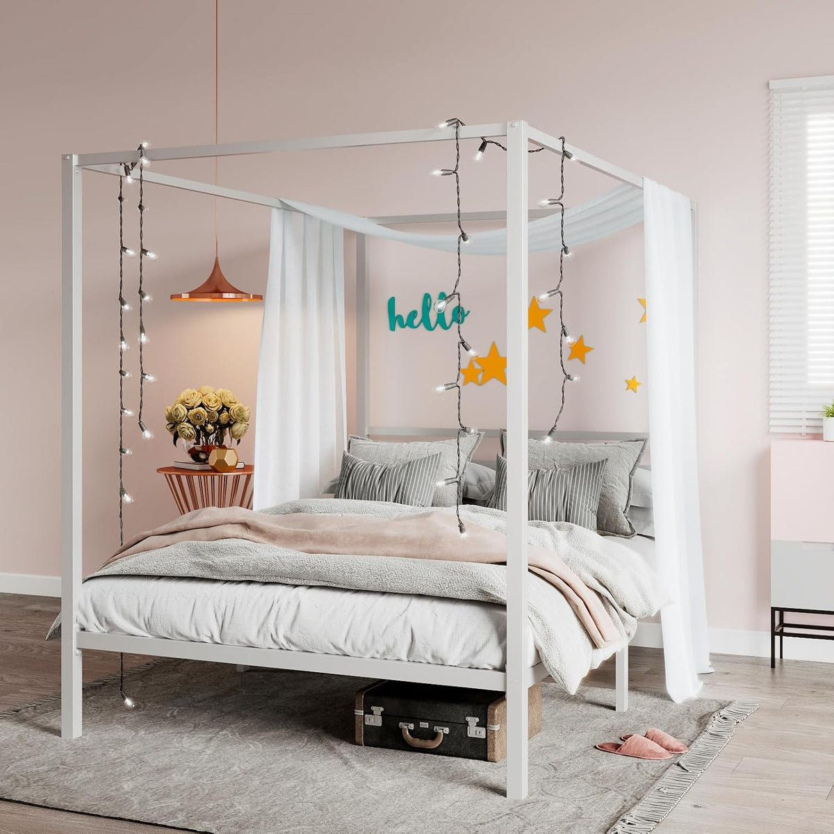 White metal four-poster canopy bed with sheer drapes, string lights, layered bedding and a suitcase under the frame.