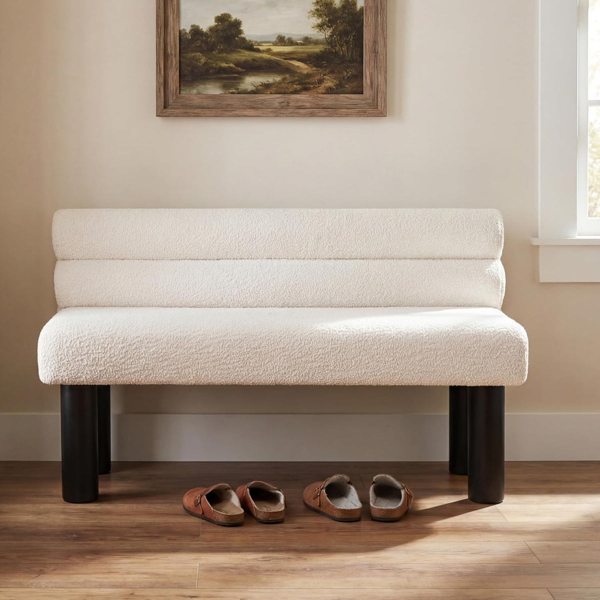 Cream boucle bench with cylindrical dark legs placed in an entryway under framed art.