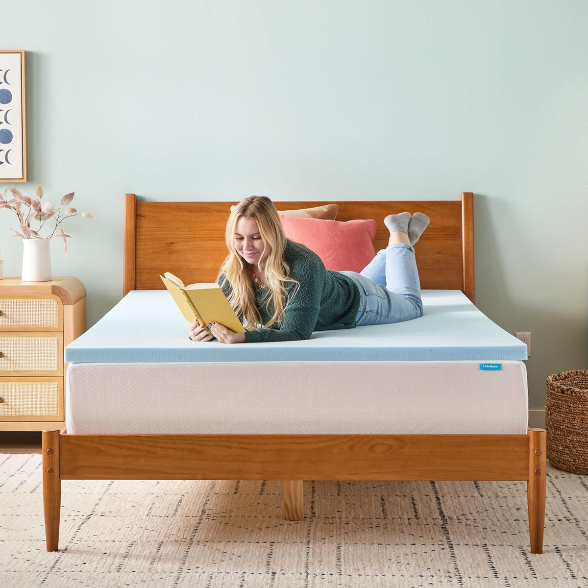 Person lying on stomach reading a book on a light-blue foam topper placed atop a mattress.