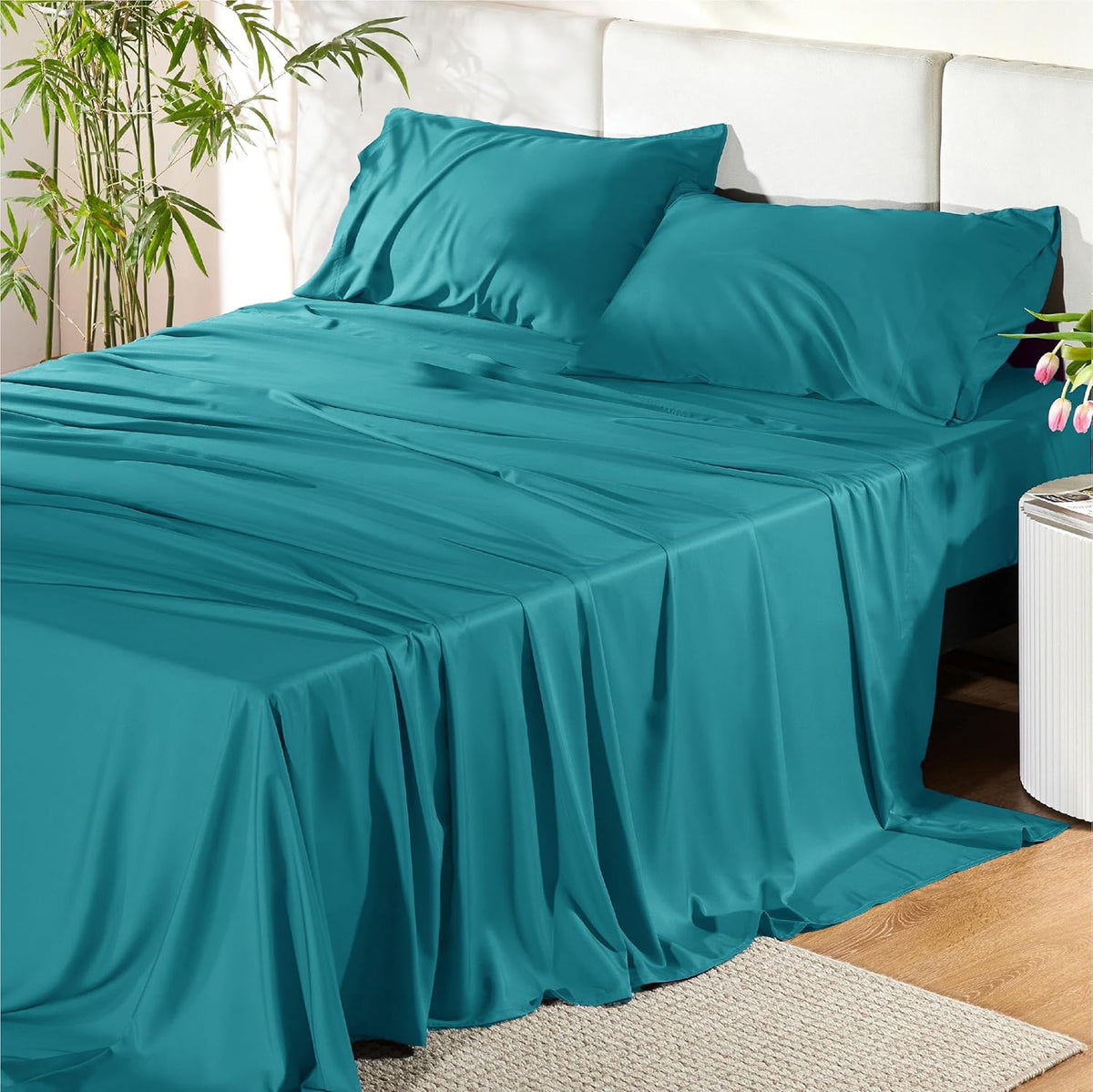 Bright teal sheet set arranged on a bed with white headboard and a small side table with flowers.
