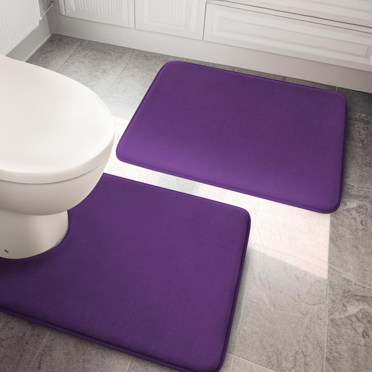 Top-down bathroom view of a purple memory-foam rug set positioned by a toilet on stone tile.