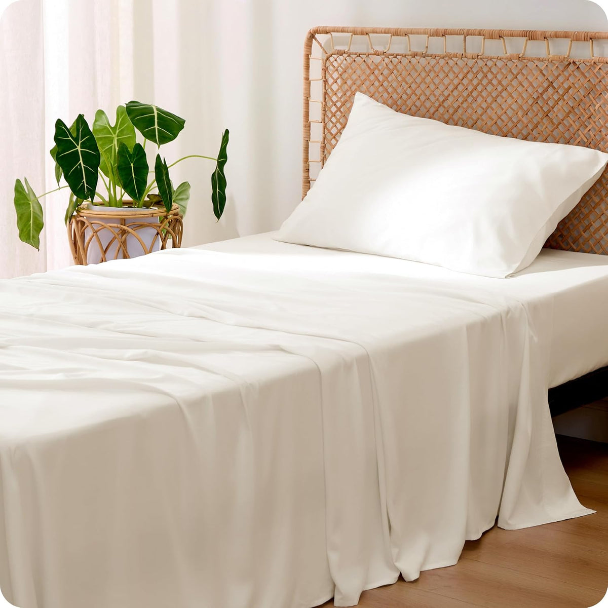 Bright bedroom scene with white silky sheets and one pillow on a bed with a woven headboard and potted plant.