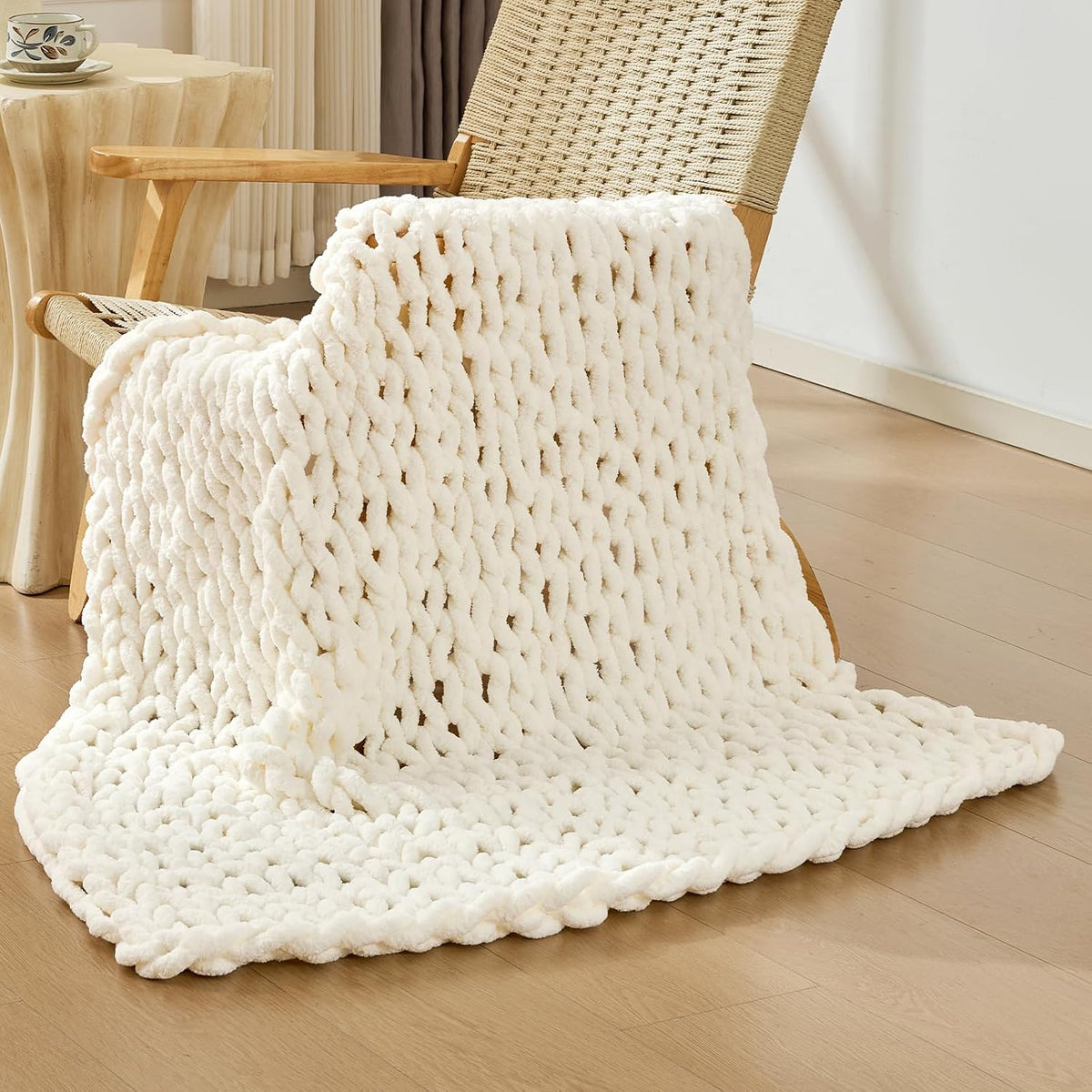 Cream-beige chunky knit throw draped over a woven-seat wooden chair, spilling onto a light wood floor.