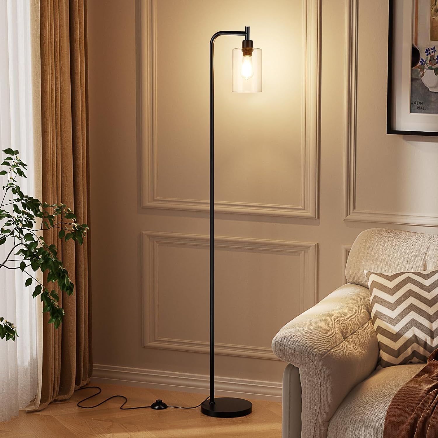 Tall black floor lamp with clear cylindrical glass shade lit beside a beige sofa; cord and foot pedal on wood floor.