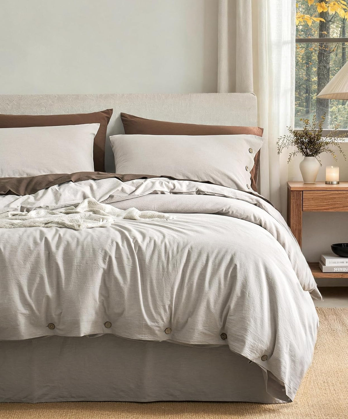 Bed styled with light gray duvet and contrasting brown pillow backs, bedside table and window at right