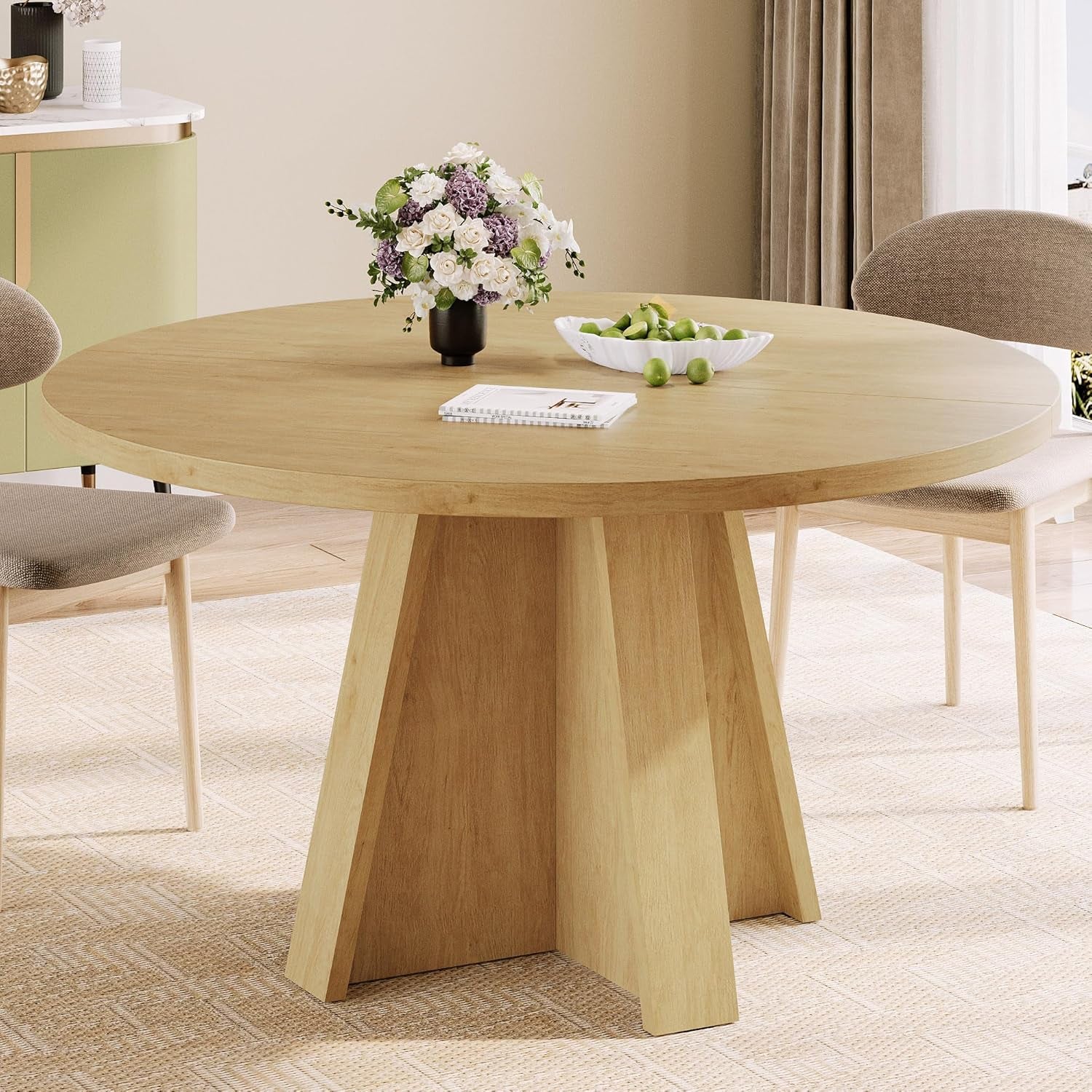 Round light-wood dining table with angular pedestal base and a bowl of green apples