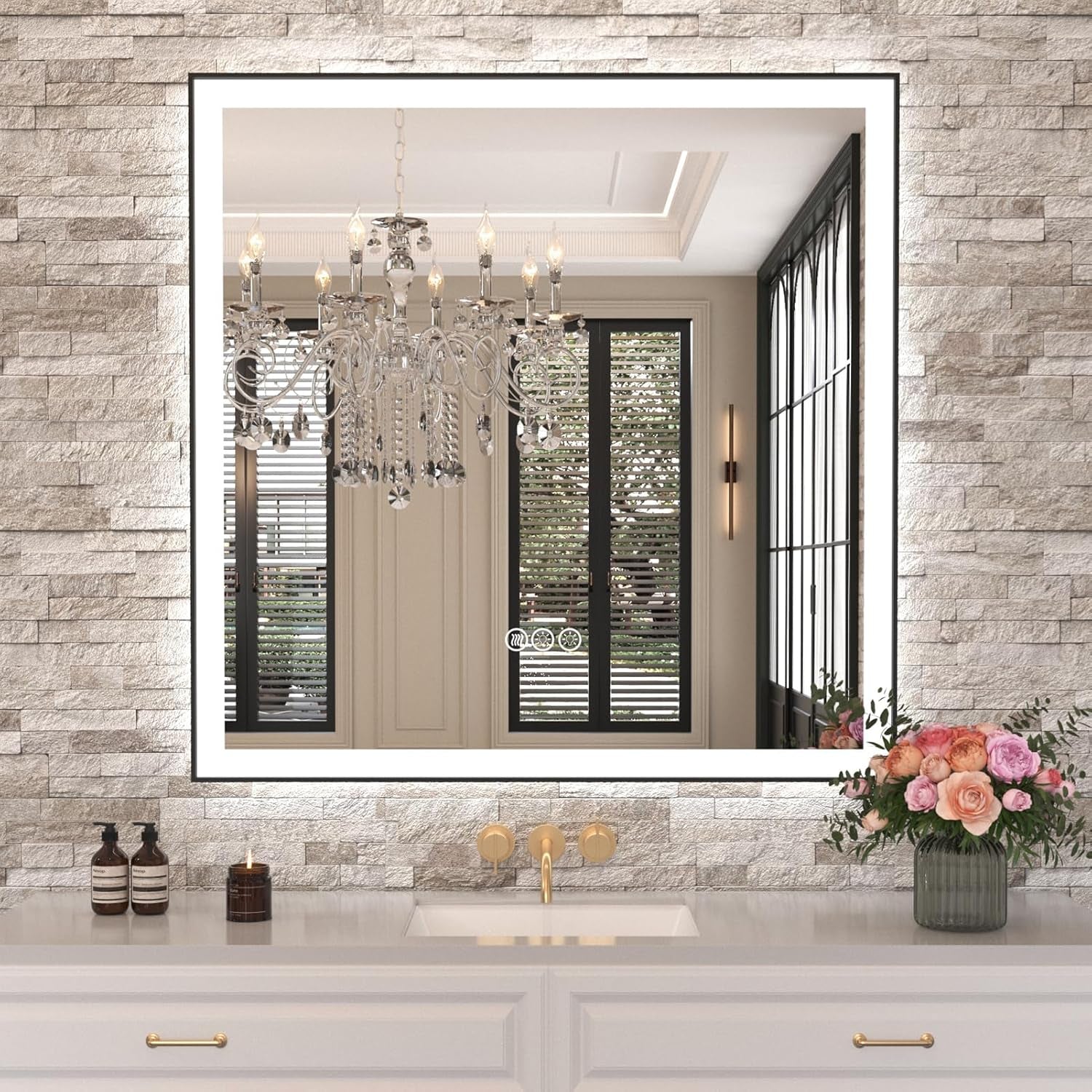 Square illuminated mirror on textured stone wall above single sink and gold faucet, vase of flowers nearby