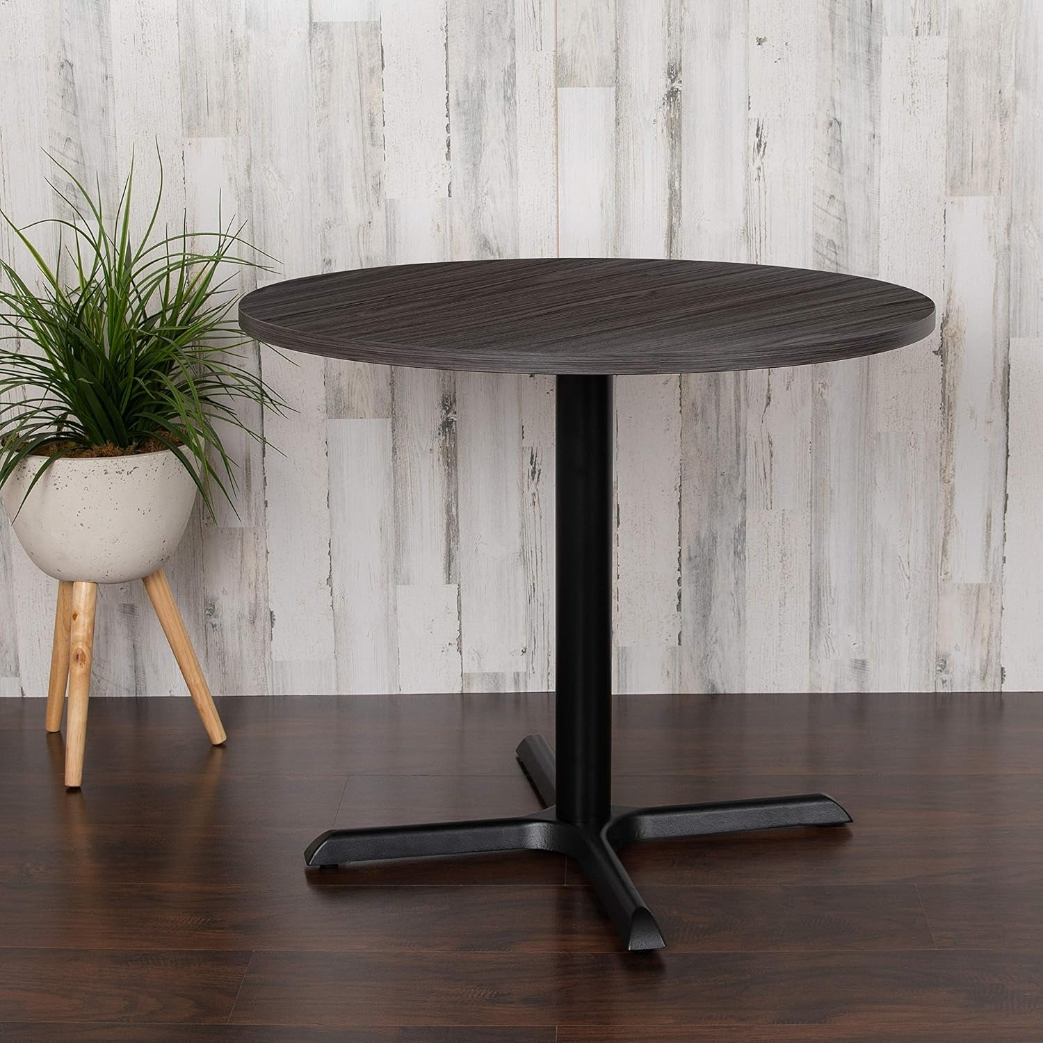 Side view of round table with dark gray woodgrain top and black X-base staged against a light wood wall.