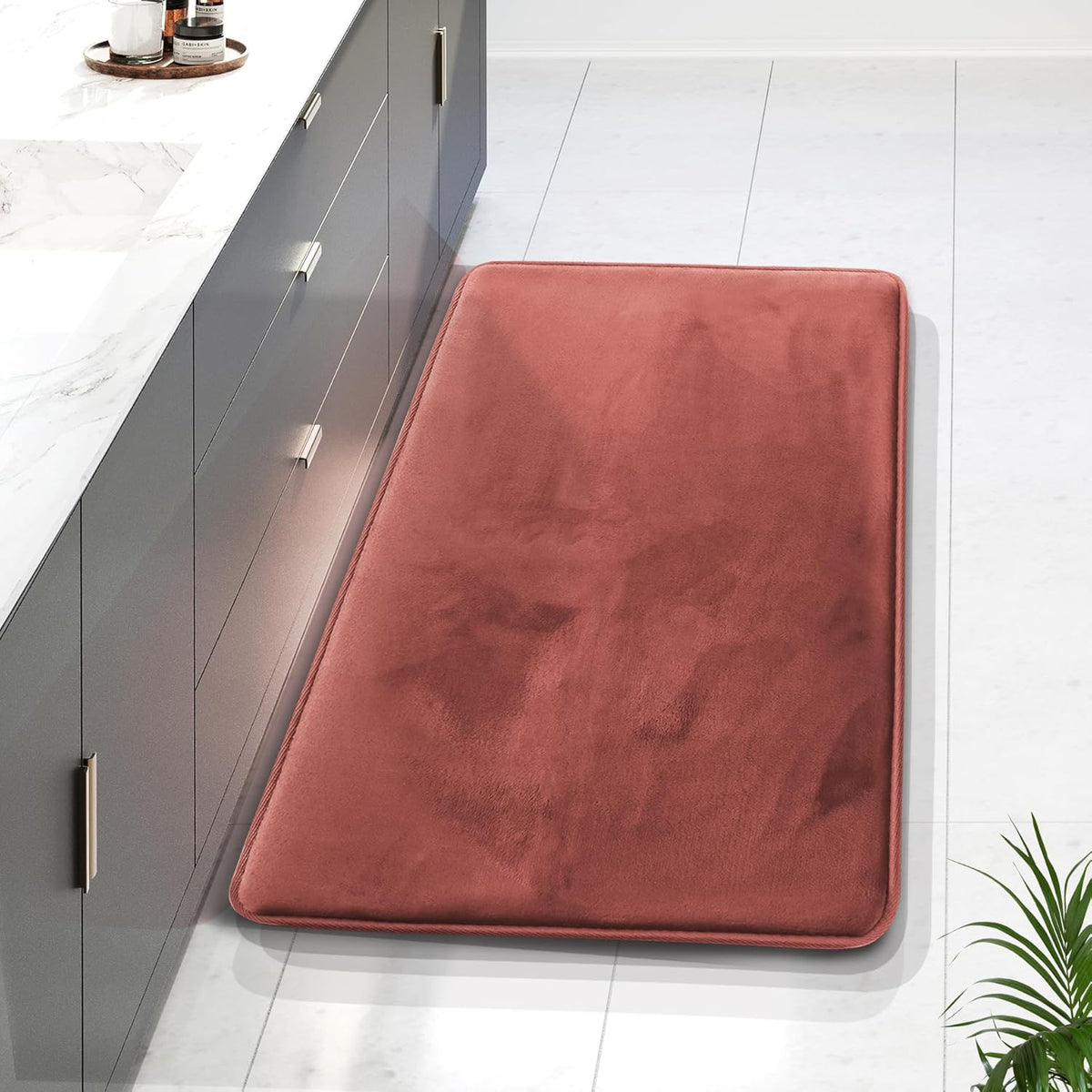 Long rust-colored rectangular bath mat beside a gray vanity on white tiled floor.