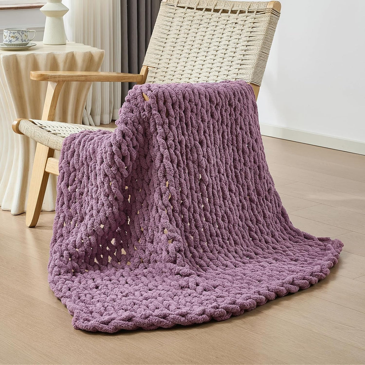 Mauve chunky knit throw draped over a woven wood chair, pooling on a light wood floor.