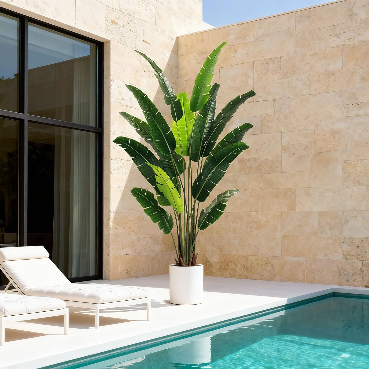 Tall artificial bird of paradise tree with large green leaves in a white round pot next to a poolside patio