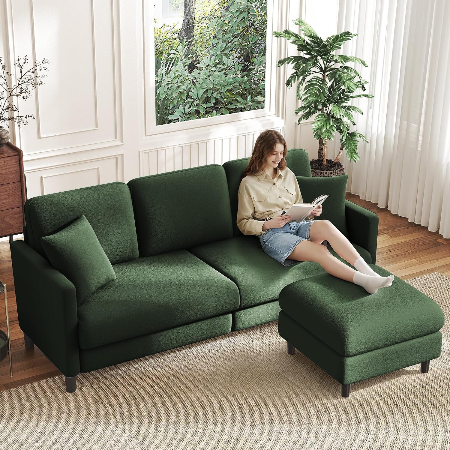 Bright room with a green three-seat sofa and matching ottoman, a woman reclining and reading a book.
