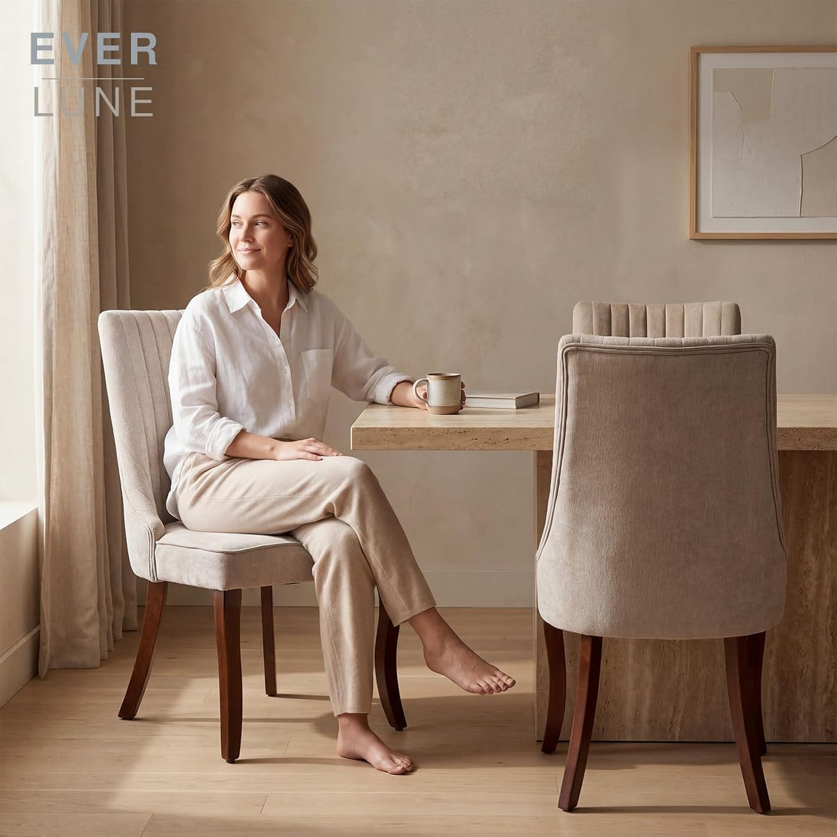 Woman seated on a beige high-back dining chair at a table with a rear view of a matching chair beside her