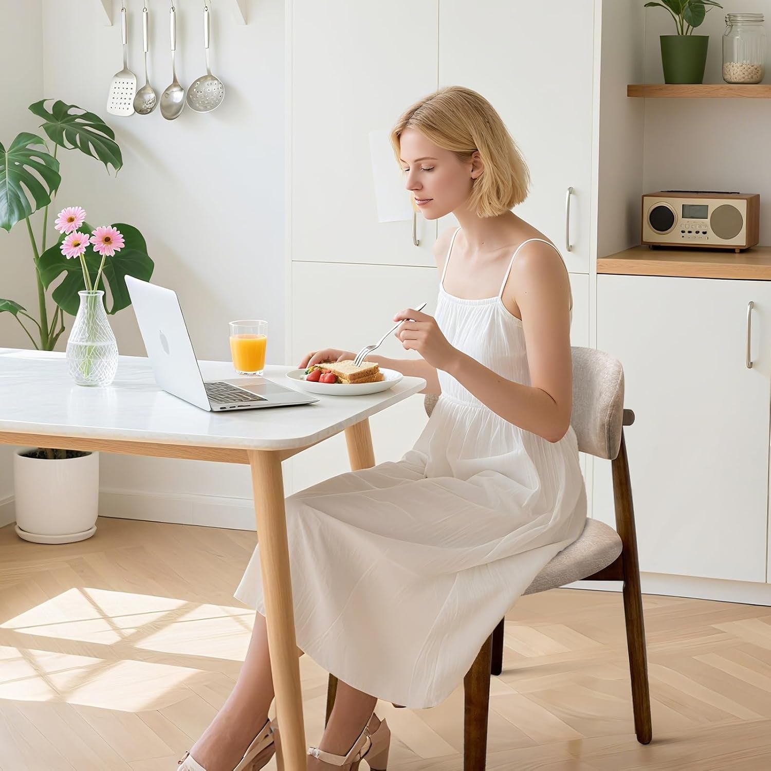 Woman sits on a beige upholstered chair while eating at a small kitchen table with a laptop.