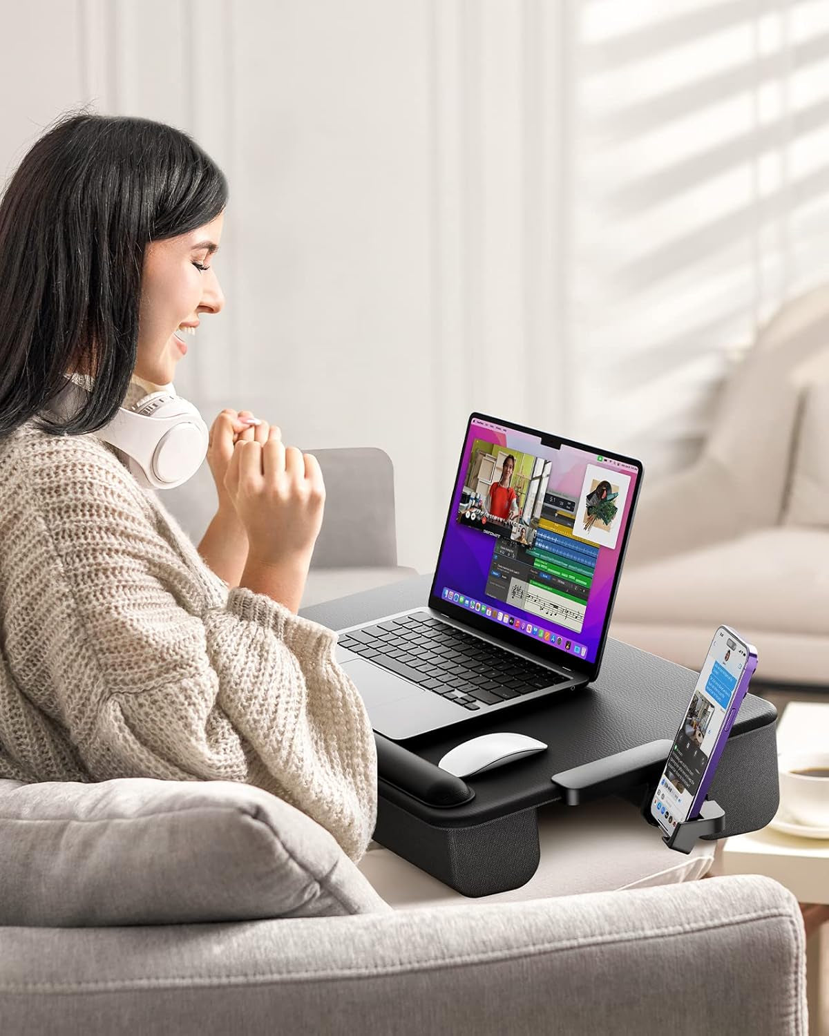 Side view of a woman sitting on a sofa using a black lap desk with a laptop, mouse and phone holder on her lap.