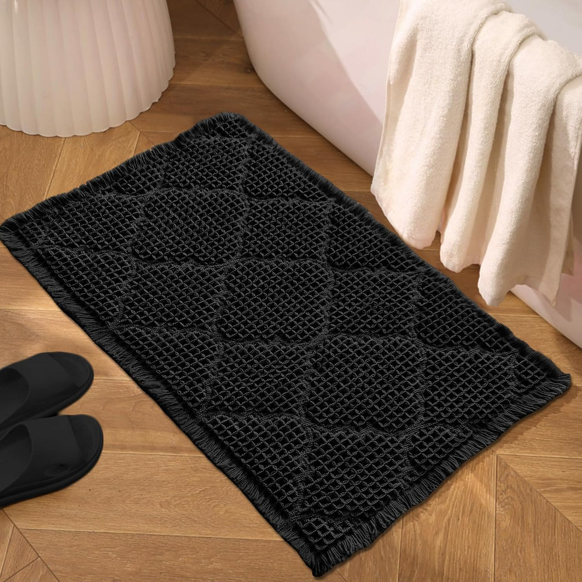 Black waffle-texture bath mat on wood floor beside bathtub and towels